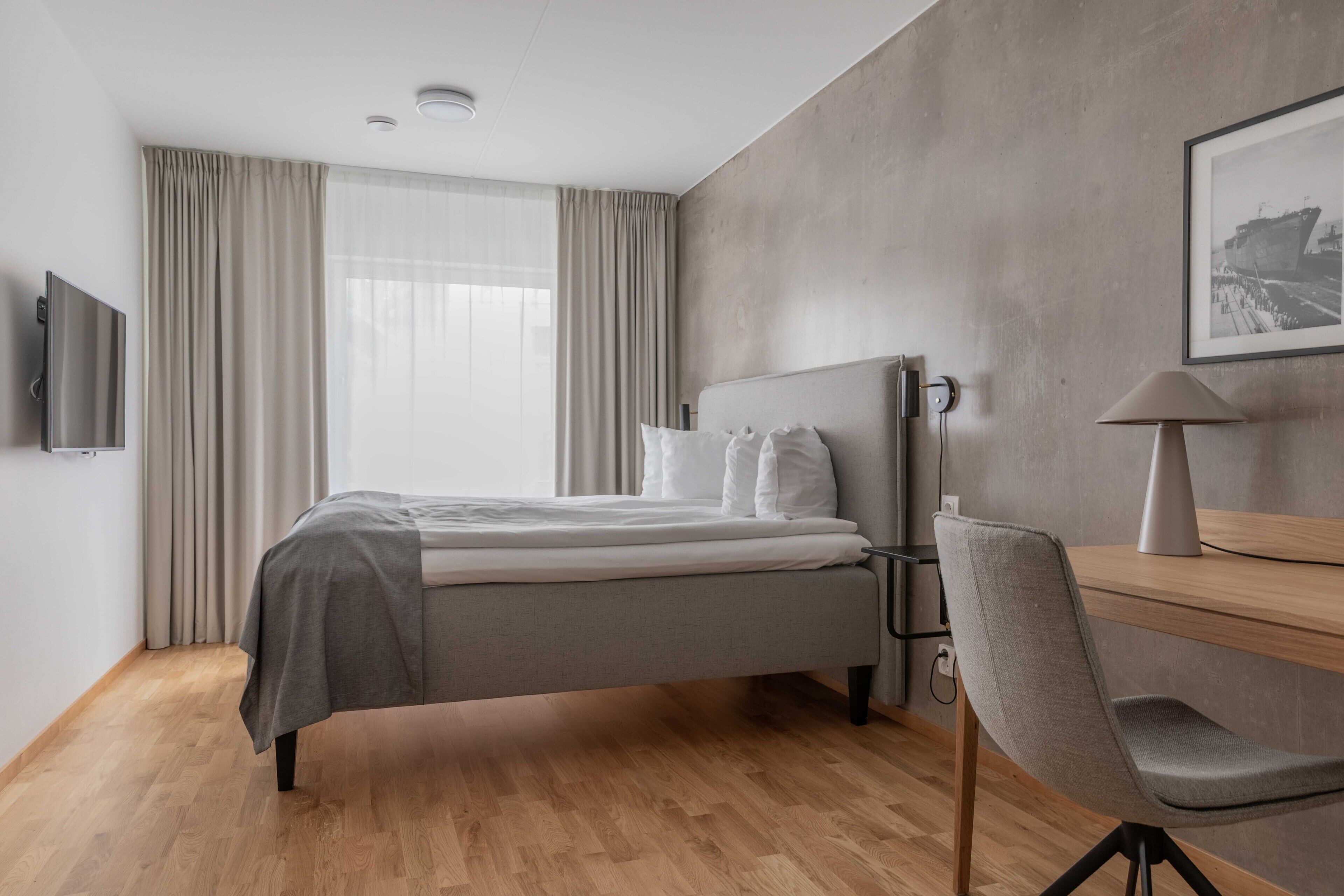 A modern hotel room with a bed, a desk, a chair, and a television.