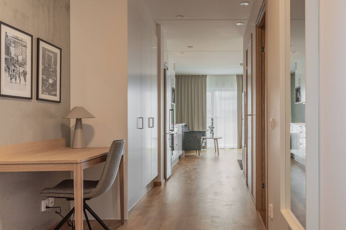 Een moderne hotelgang met een houten bureau, een stoel, afbeeldingen in lijsten aan de muur, en uitzicht op een kamer met een bed en een raam.