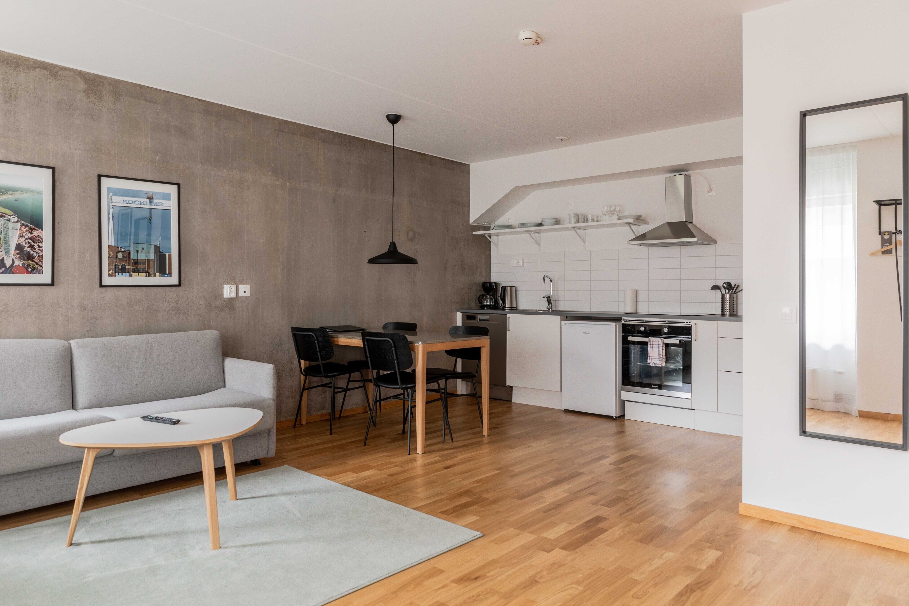 Een moderne woonkamer met een bank, een eettafel met stoelen, een koffieapparaat en een keuken met witte kasten en een fornuis.