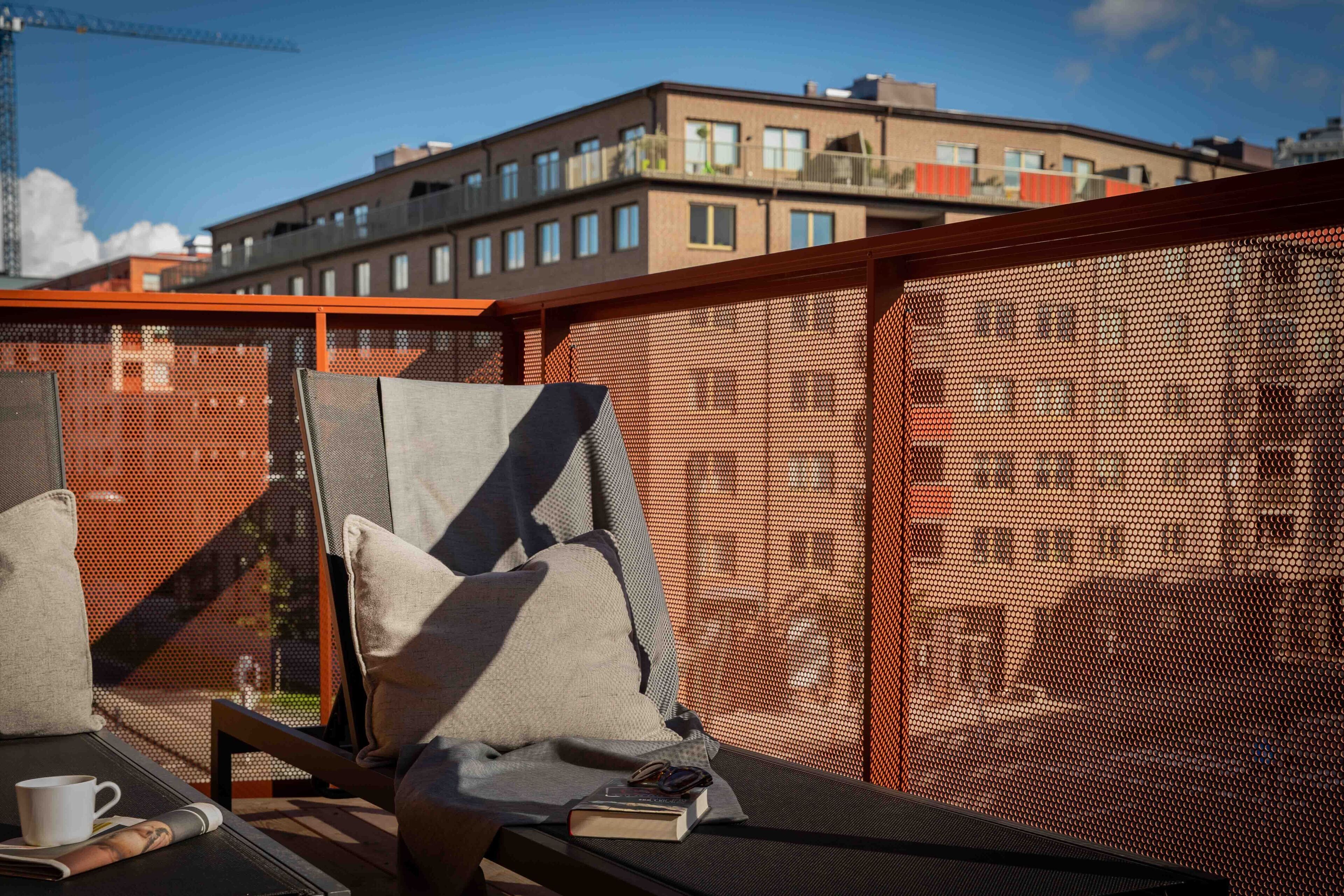 A balcony with a mesh screen, a chair, a cushion, a table with a cup and a book, and a view of a building in the background.