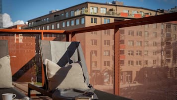 Ein Balkon mit einem Insektenschutznetz, einem Stuhl, einem Kissen, einem Tisch mit Tasse und Buch, und Blick auf ein Gebäude im Hintergrund.