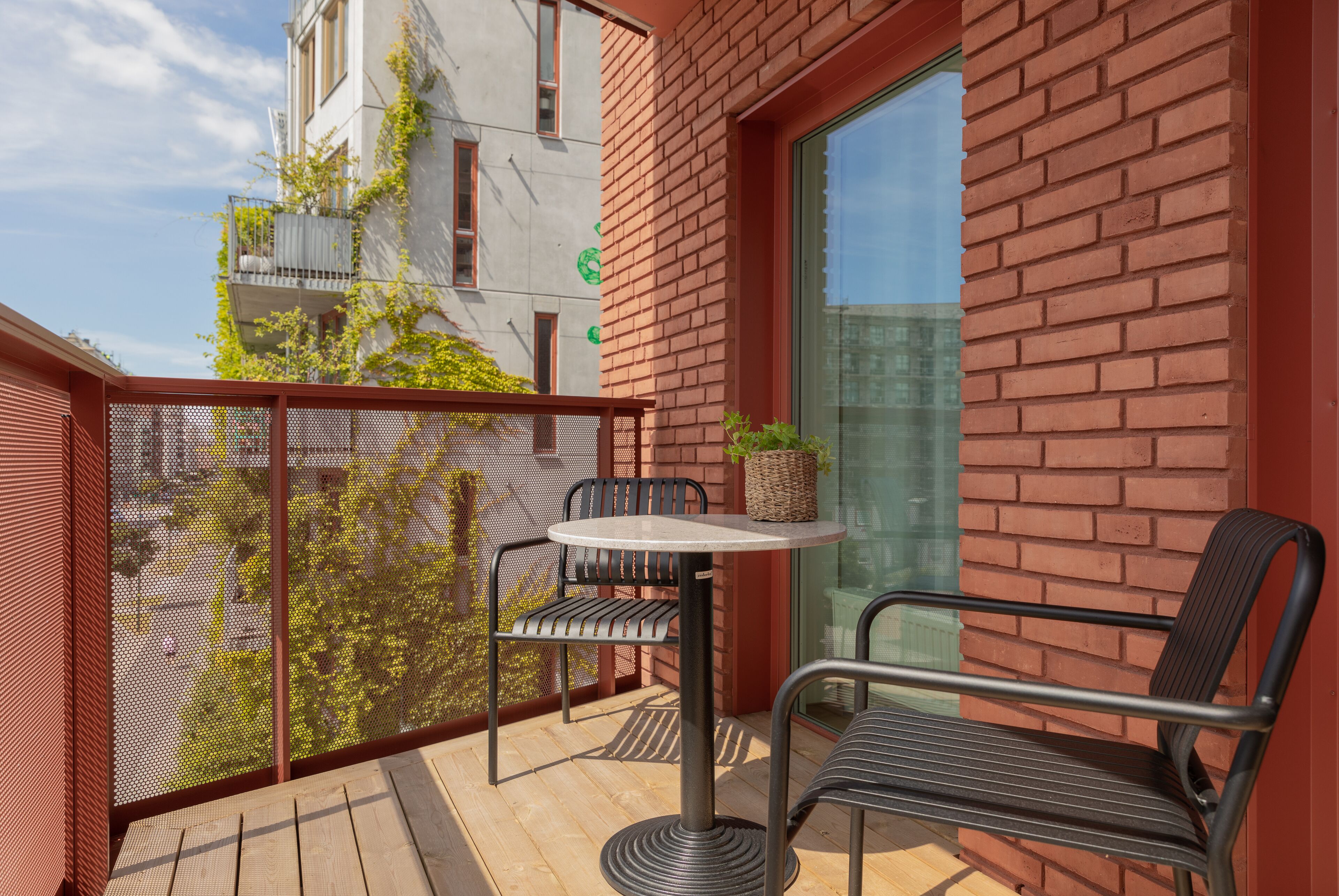 A balcony with a round table and two chairs, a red brick wall, and a view of a building with a balcony.