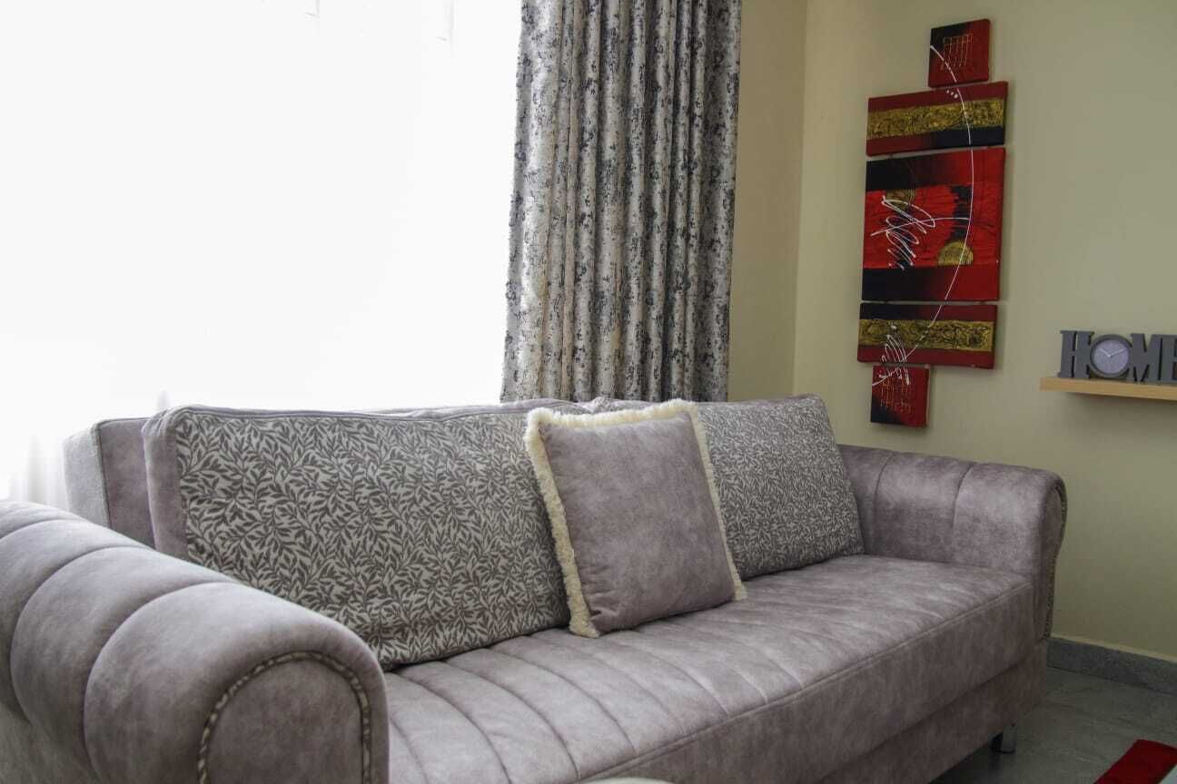 A grey sofa with patterned cushions, a window with sheer curtains, and a wall with abstract art and a sign.