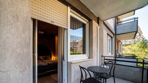 Une porte coulissante ouverte donne sur une pièce où se trouvent un lit et un mur éclairé. Dehors, il y a un balcon avec une table et des chaises.