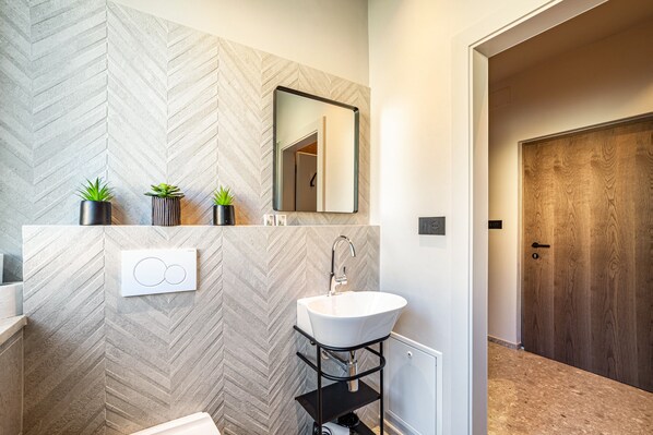 Une salle de bain moderne avec un mur orné d’un motif en chevrons, un lavabo blanc, un miroir et trois plantes en pot.
