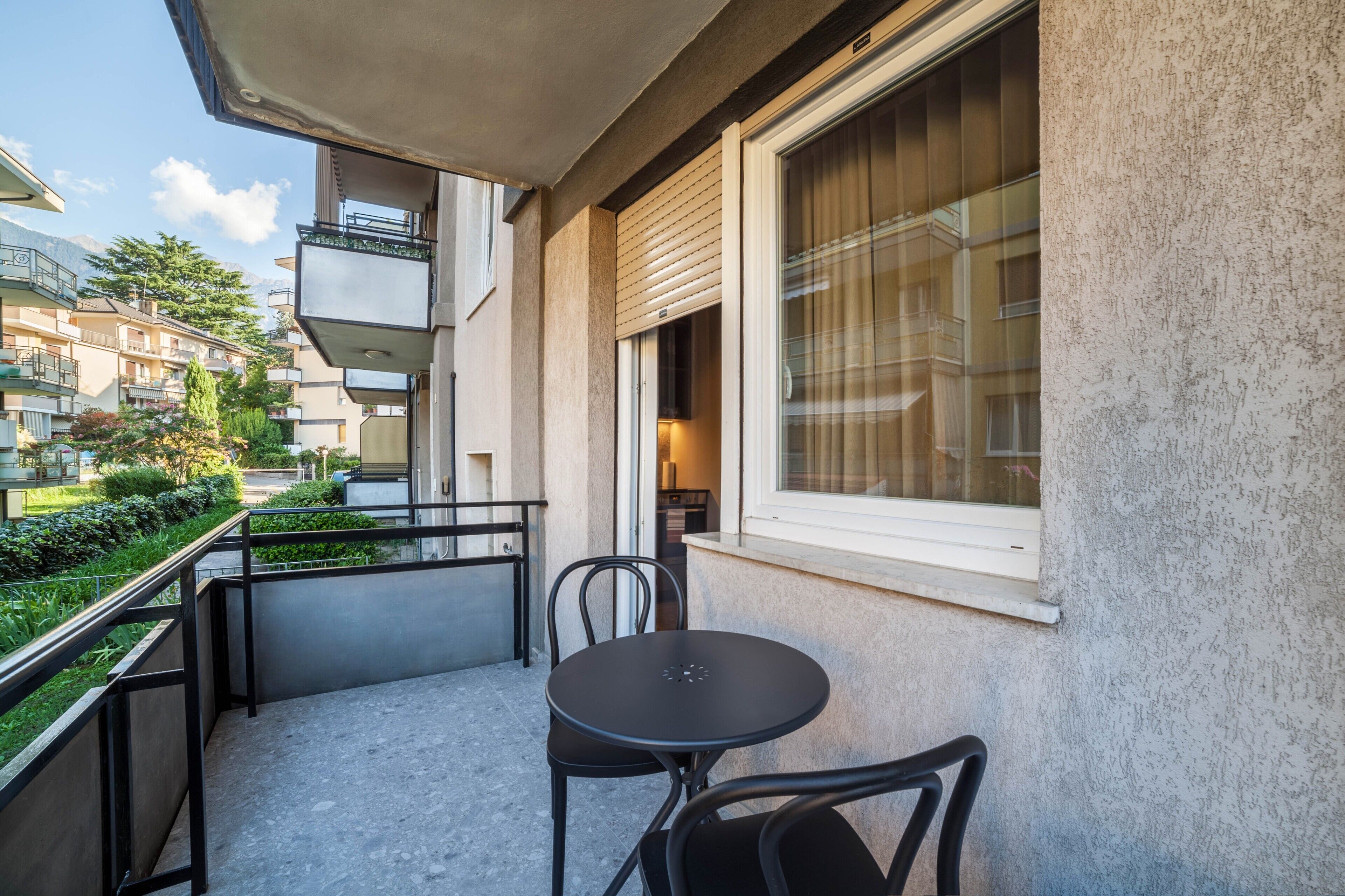 Un balcon avec une table et des chaises, une fenêtre avec des stores et une vue sur des bâtiments et de la verdure.