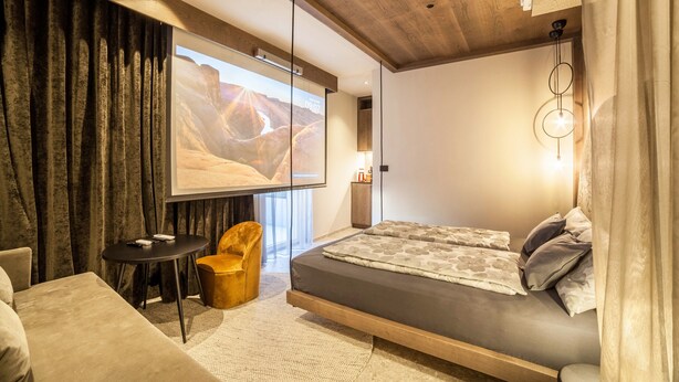 Une chambre moderne avec un lit, une petite table, une chaise et un grand écran affichant un paysage désertique.