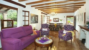 Un salon avec un canapé violet, des fauteuils à motifs, une table basse et un coin repas agrémenté d’un lustre.