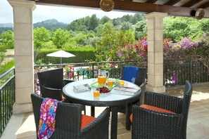 Ein überdachter Außenbereich mit Korbstühlen, einem Tisch mit Getränk und Blick auf einen üppigen Garten.