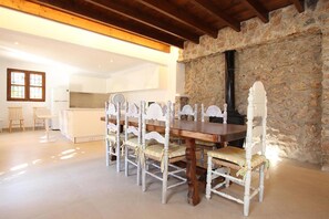A dining area with a wooden table and white chairs, a stone wall, and a kitchen in the background.