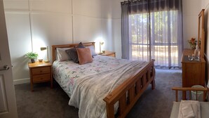 A bedroom with a wooden bed, a nightstand with a lamp, a small table with a vase, and a window with sheer curtains. - Loves Road Retreat   (Barwon Downs)