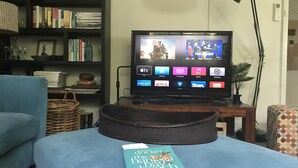 A living room with a television displaying various streaming services, a bookshelf, a lamp, and a book on a table. - Loves Road Retreat   (Barwon Downs)