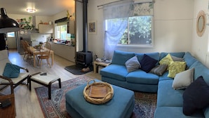 A cozy living room with a blue sofa, a dining area with wooden chairs, a kitchen with a stove, and a large window with curtains. - Loves Road Retreat   (Barwon Downs)