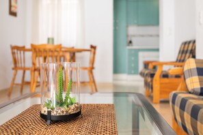 A clear glass terrarium with plants on a woven placemat, a dining table with chairs, and a sofa with plaid cushions in the background.