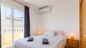 Een slaapkamer met een bed, twee bedlampen, een airconditioner, een raam met gordijnen en uitzicht op een balkon.