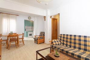 A living room with a sofa, a dining table with chairs, a kitchen area, and a clock on the wall.