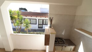 Une petite cuisine équipée d’une cuisinière à gaz, une fenêtre donnant sur un immeuble avec un toit en tuiles, et un balcon agrémenté de plantes.