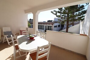 Un balcon avec une table et des chaises blanches, un pot de fleurs et une vue sur des immeubles et un arbre.