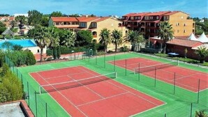 A tennis court surrounded by buildings with red roofs and a pool in the background.
