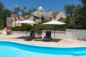 Un espace piscine avec des chaises longues et des parasols, un bâtiment en pierre en arrière-plan et un ciel bleu dégagé.