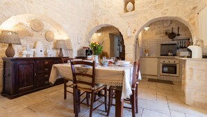 A rustic stone kitchen with a dining table set for four, wooden chairs, and a stone oven. - Holiday Home Trullo Nurillo with Pool & Wi-Fi (Alberobello)