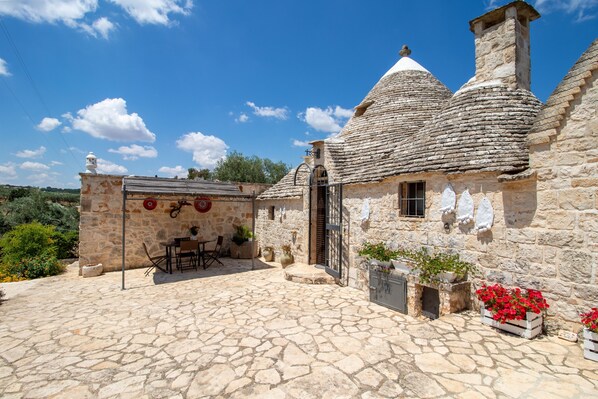 Una casa de piedra con tejado de baldosas, un pequeño patio con mesa y sillas, y plantas en macetas.