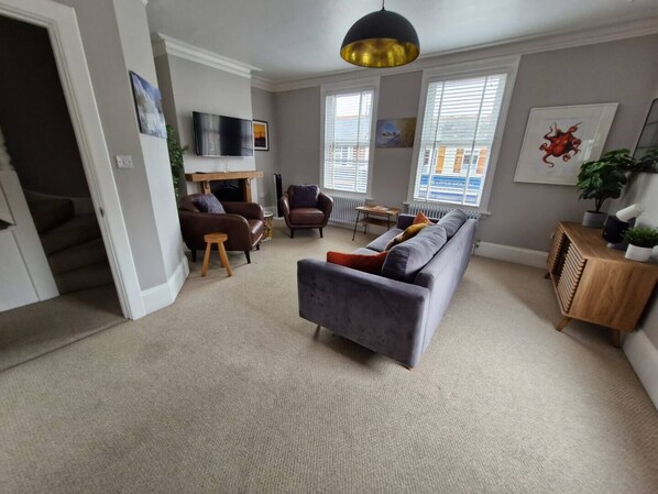 A modern living room with a grey sofa, two armchairs, a coffee table, and a TV mounted on the wall. There is a fireplace, a wooden sideboard, and a large window with blinds. - The Old Chemist - Viking Bay Getaway - Mins To Beach and Town (Kent)