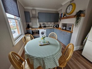 A compact kitchen with a dining area, featuring a table with a checkered tablecloth, wooden chairs, a washing machine, and a microwave. - The Old Chemist - Viking Bay Getaway - Mins To Beach and Town (Kent)