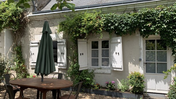 A patio with a round wooden table and chairs, a green umbrella, and a white building with shutters and a door. - Les Hortensias, 3-star self-catering cottage, heated pool, wifi, games... (Monteaux)