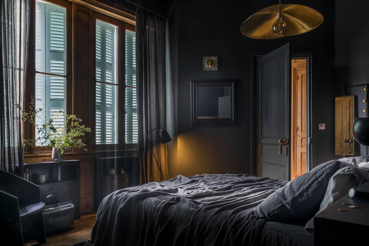 Ein Schlafzimmer mit einem großen Bett, einem Fenster mit Fensterläden, einem Schreibtisch mit Lampe und einer Tür zu einem anderen Raum.