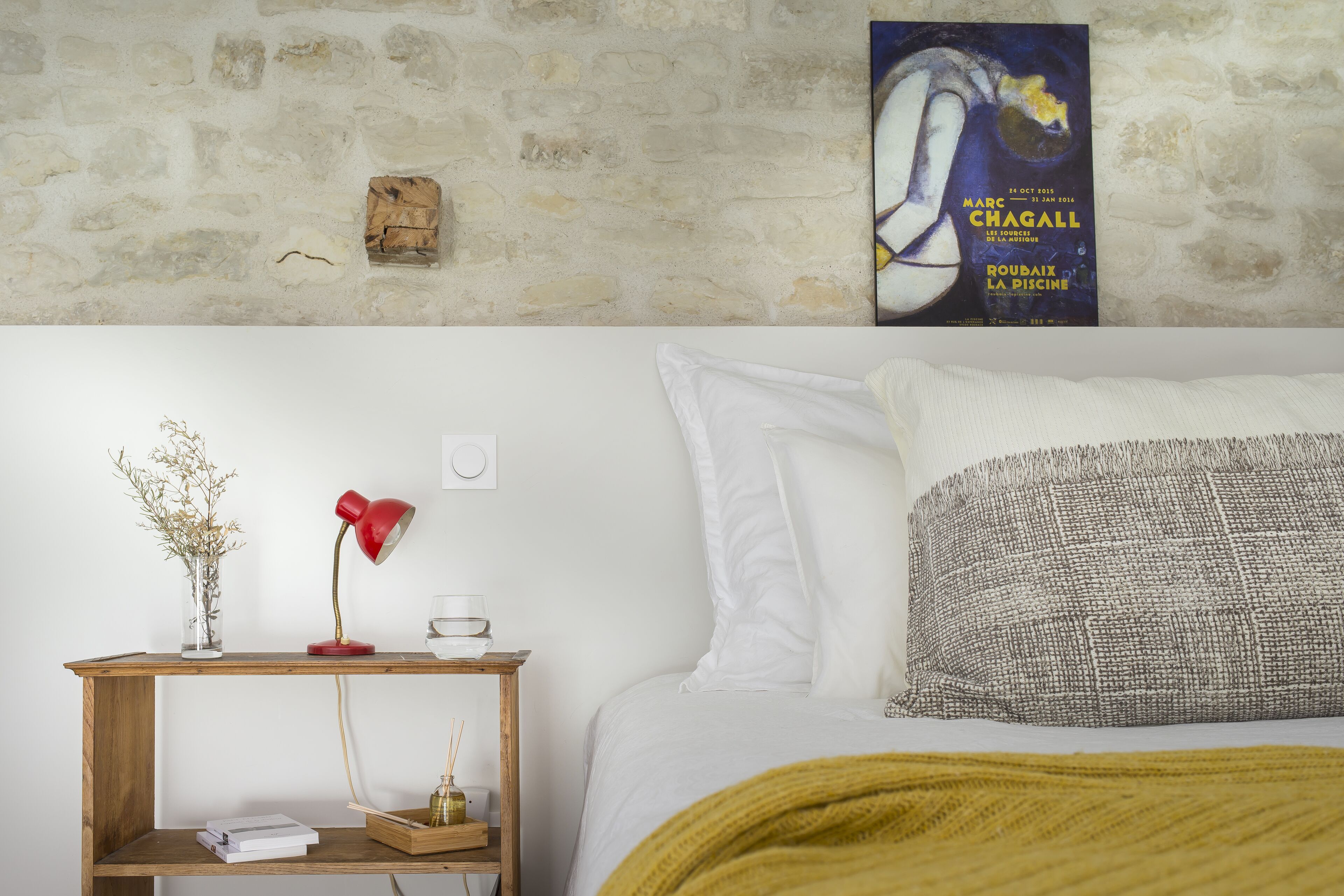 A bedroom with a bed, a nightstand with a lamp, a framed poster, and a stone wall.