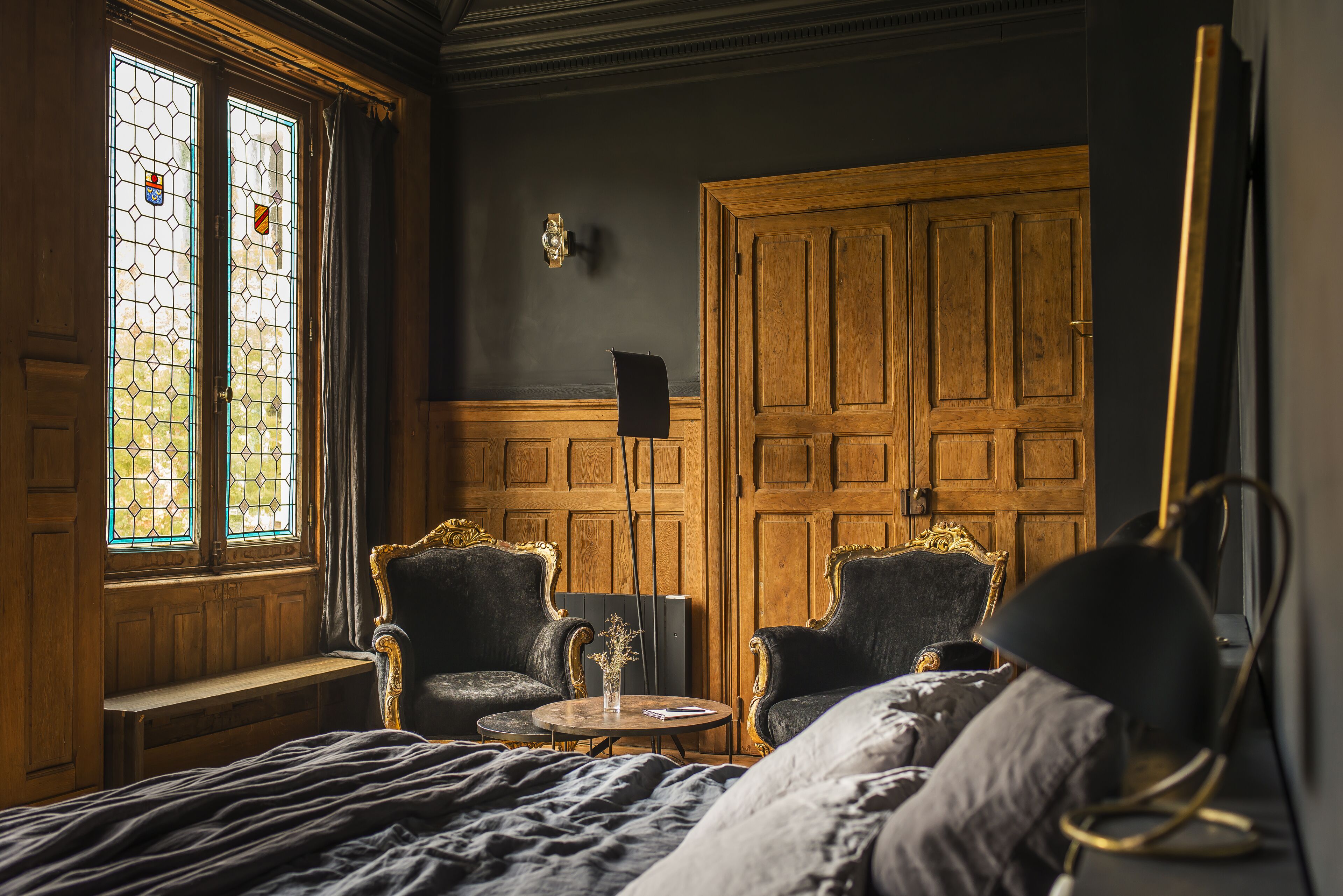 A room with a large wooden wardrobe, two armchairs, a round table with a vase, and a bench.