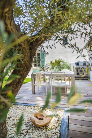 Un patio al aire libre con mesa y sillas de madera, rodeado de follaje y una pared blanca.