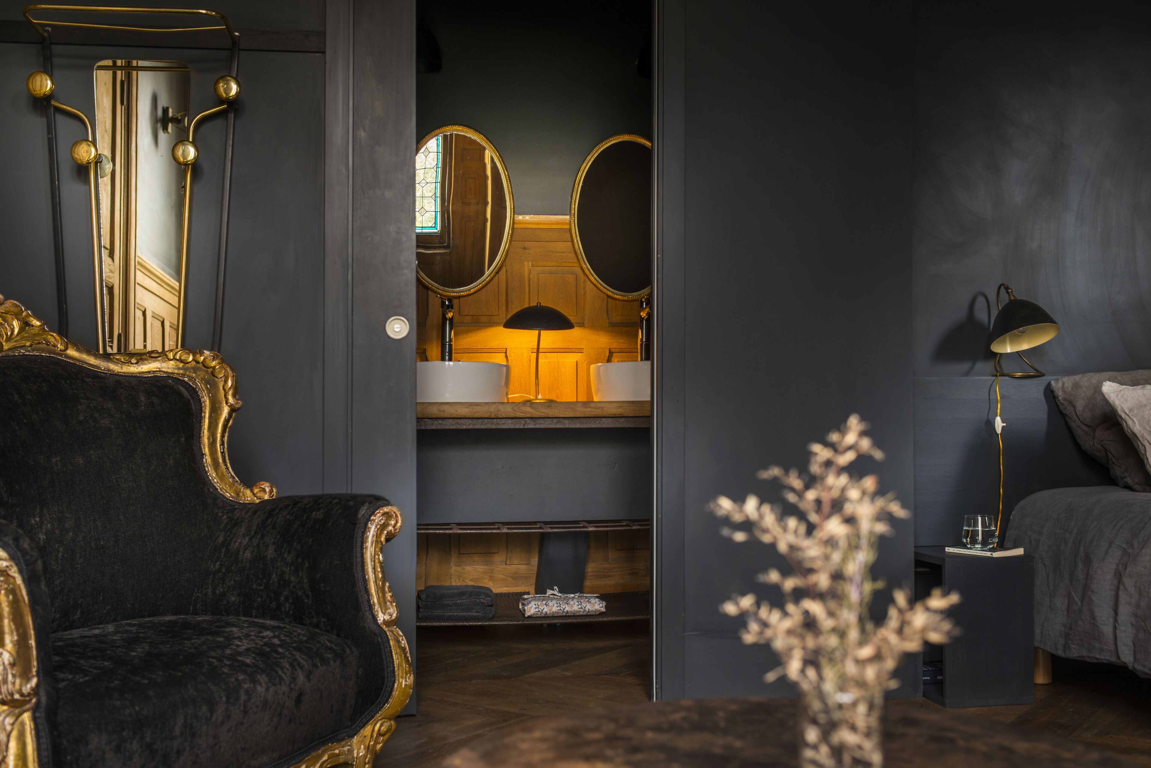Une pièce aux couleurs sombres, comprenant un fauteuil noir capitonné, une table de chevet avec une lampe et un miroir au-dessus des toilettes.
