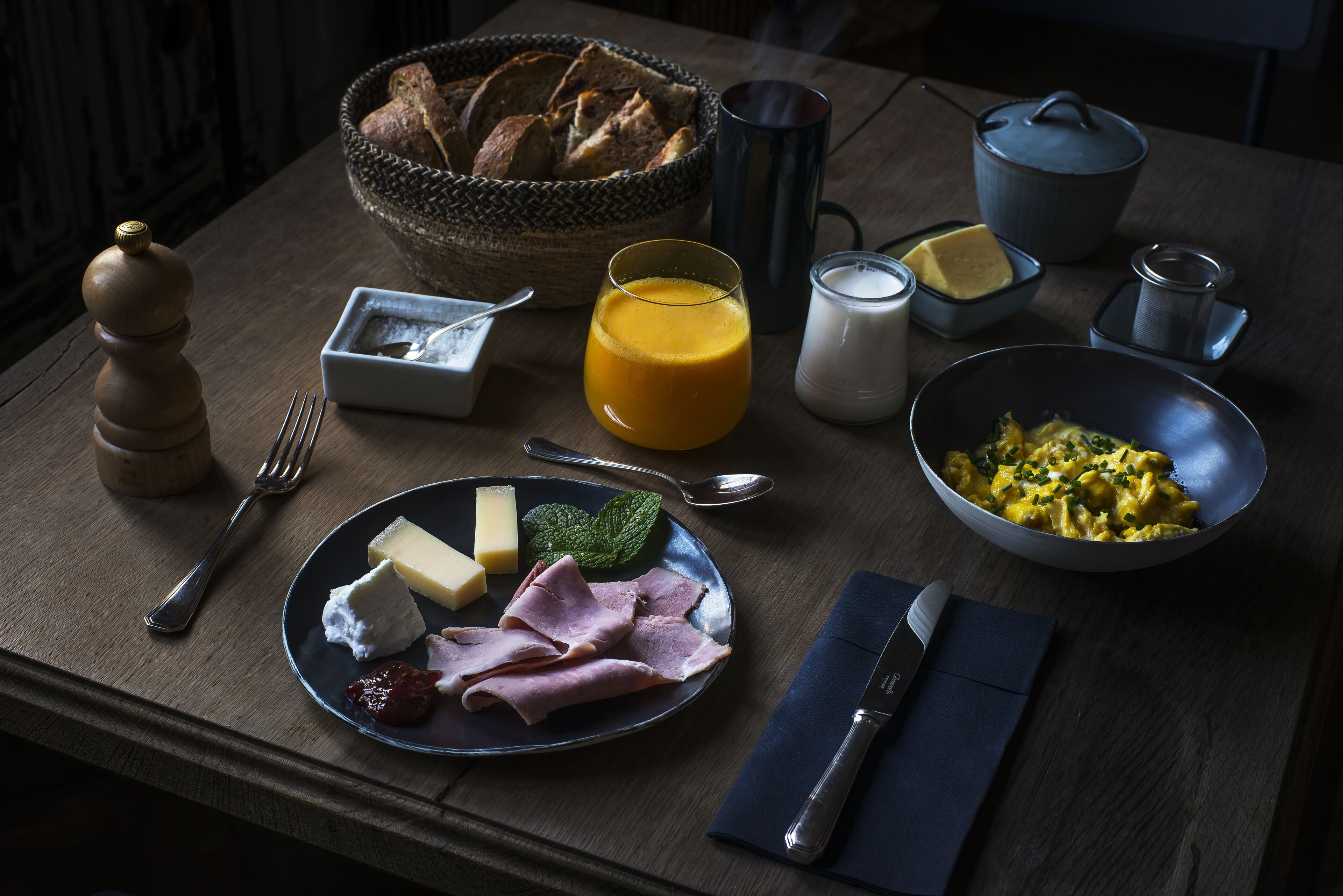 Uma mesa de café da manhã com pão, queijo, presunto e um recipiente com ovos mexidos.