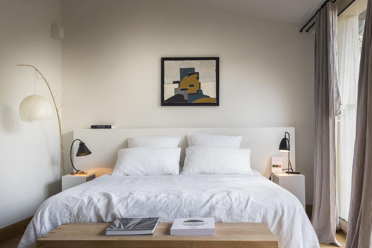 Ein ordentlich eingerichtetes Schlafzimmer mit einem großen Bett, zwei Nachttischen, einer Nachttischlampe, einer Stehlampe und einem gerahmten Kunstwerk an der Wand.