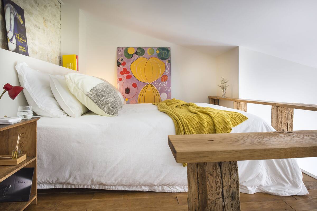 Ein Schlafzimmer mit einem hölzernen Bett, weißer Bettwäsche, einer gelben Tagesdecke, einem Holz-Nachttisch, einer roten Lampe und einem farbenfrohen abstrakten Gemälde an der Wand.