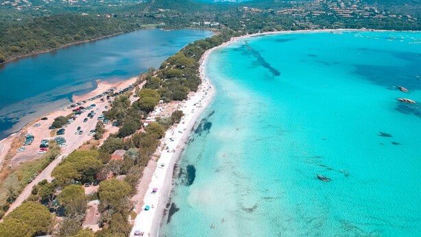 Vista aérea de uma área costeira com uma extensa praia de areia, águas turquesa cristalinas e montanhas ao fundo.