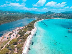 Une vue aérienne d’une zone côtière avec une longue plage de sable, des eaux turquoise et limpides, et un paysage en arrière-plan composé de collines et de montagnes.