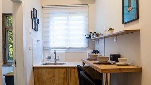 A compact kitchen with a sink, wooden cabinets, a window with blinds, a small dining table with chairs, and a shelf with kitchenware. - Aju Ben Yehuda 23 (Jerusalem)