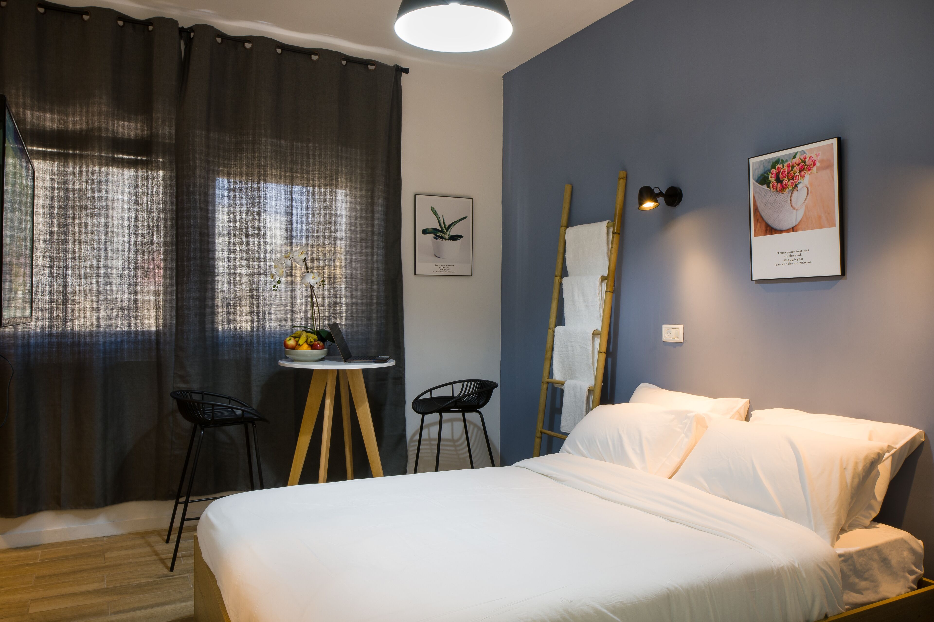 A hotel room with a bed, a desk with a chair, a small table with a bowl of fruit, and a wall-mounted lamp.