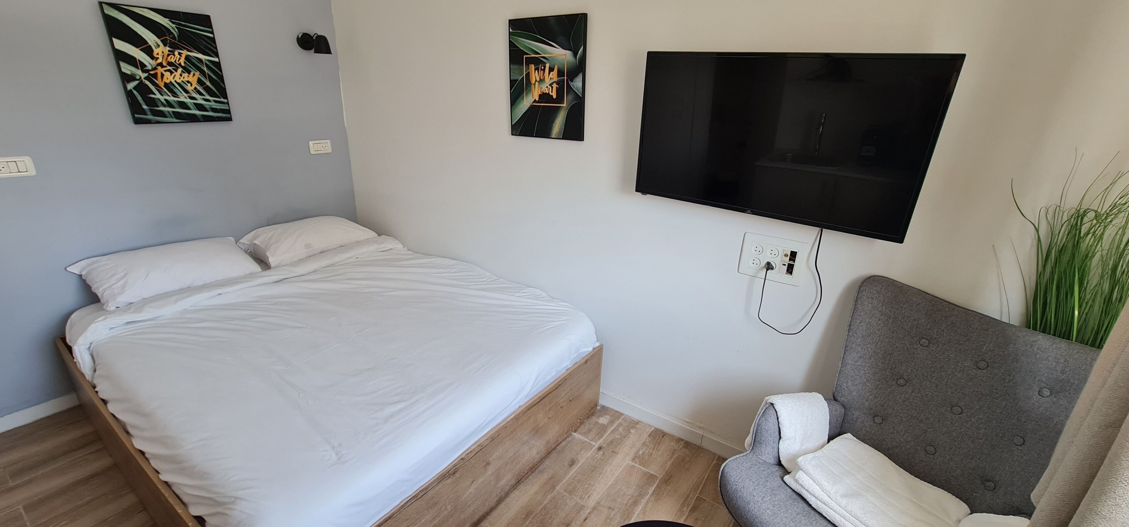 A bedroom with a bed, a gray armchair, a TV mounted on the wall, and two wall art pieces.