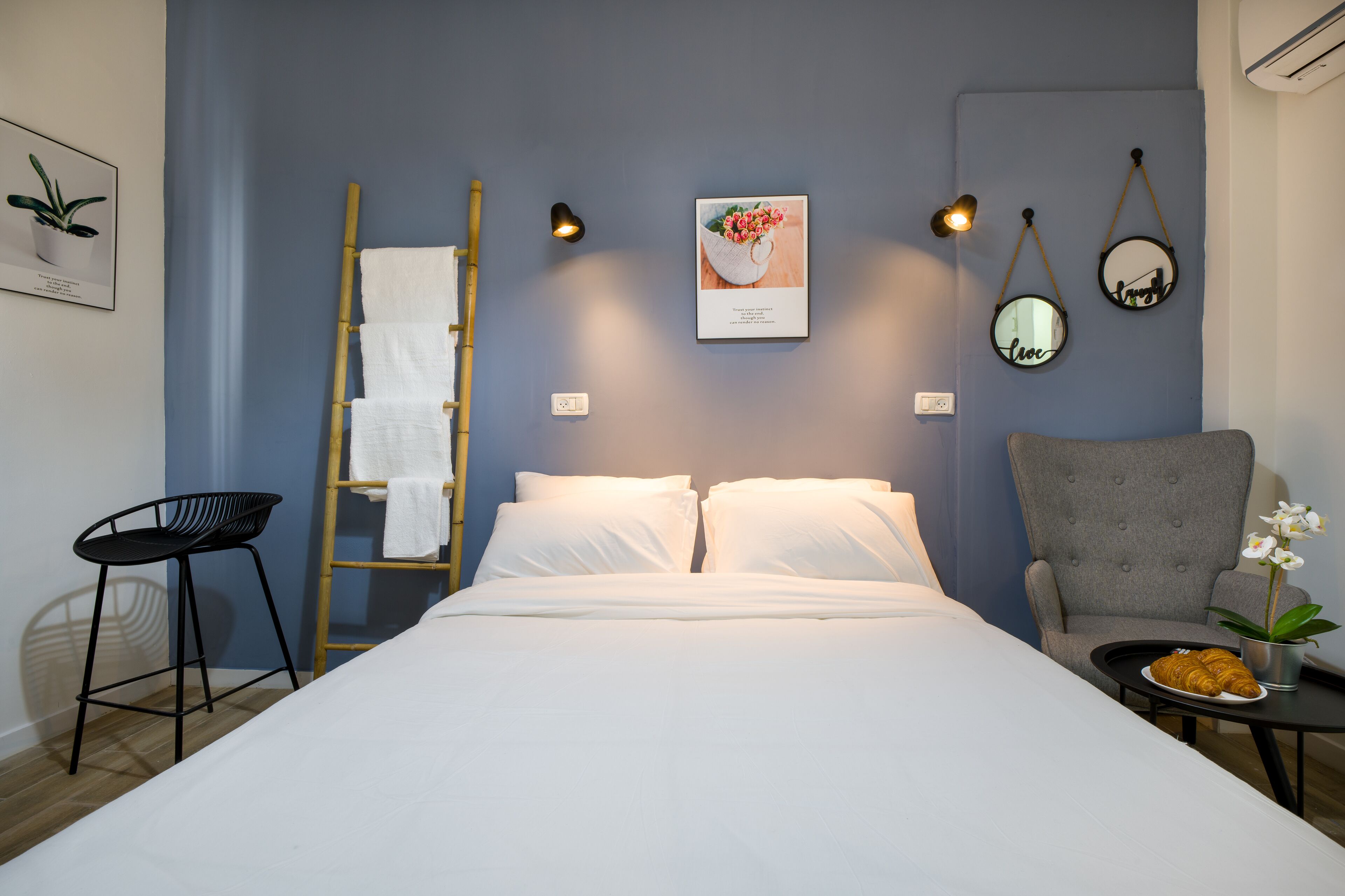 A neatly made bed with white linens, a gray armchair, a small table with a plate of croissants, and wall-mounted decor including a framed picture and two mirrors.