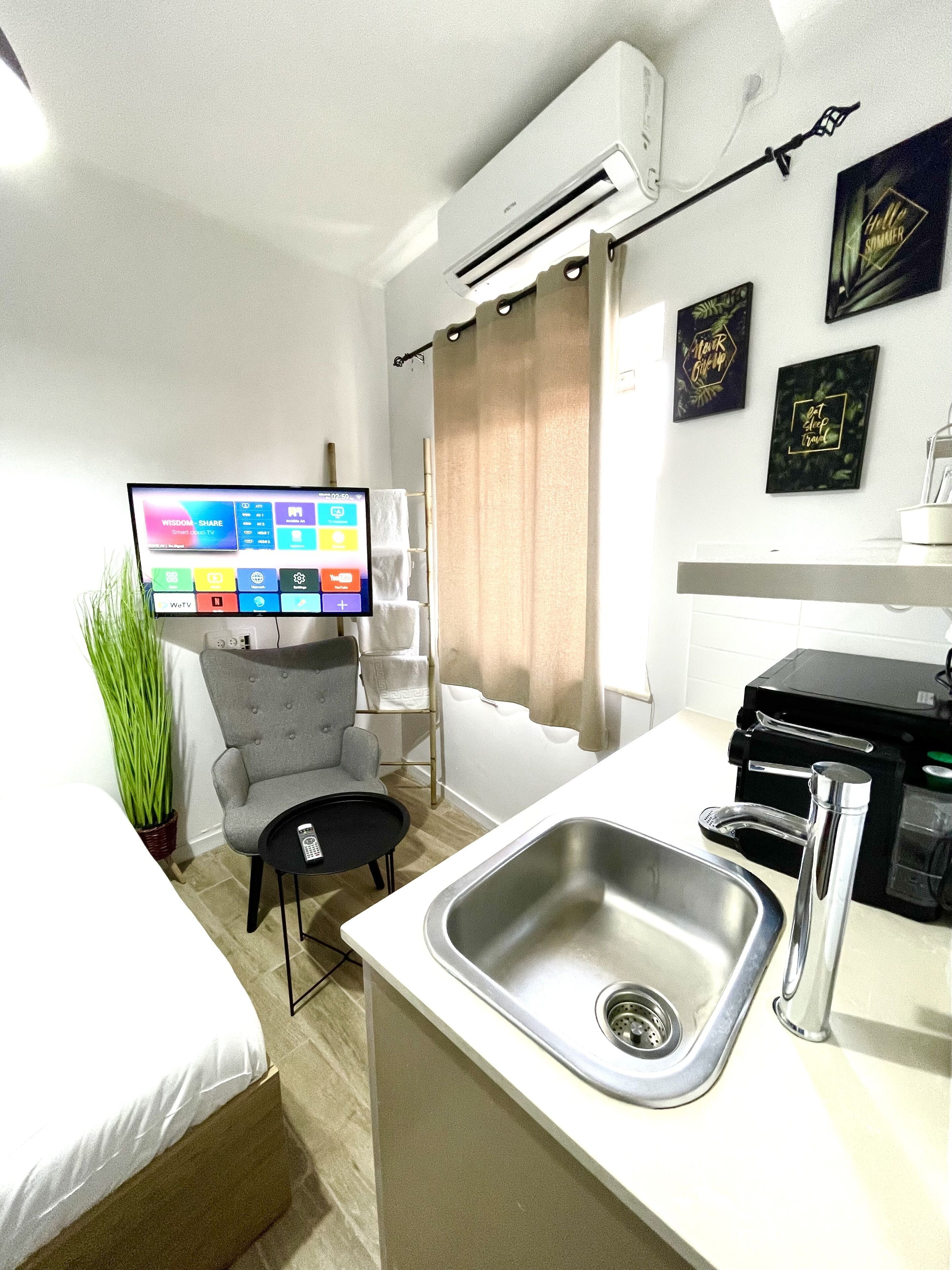 A modern hotel room with a bed, a small desk area with a computer, a sink, and a window with curtains.