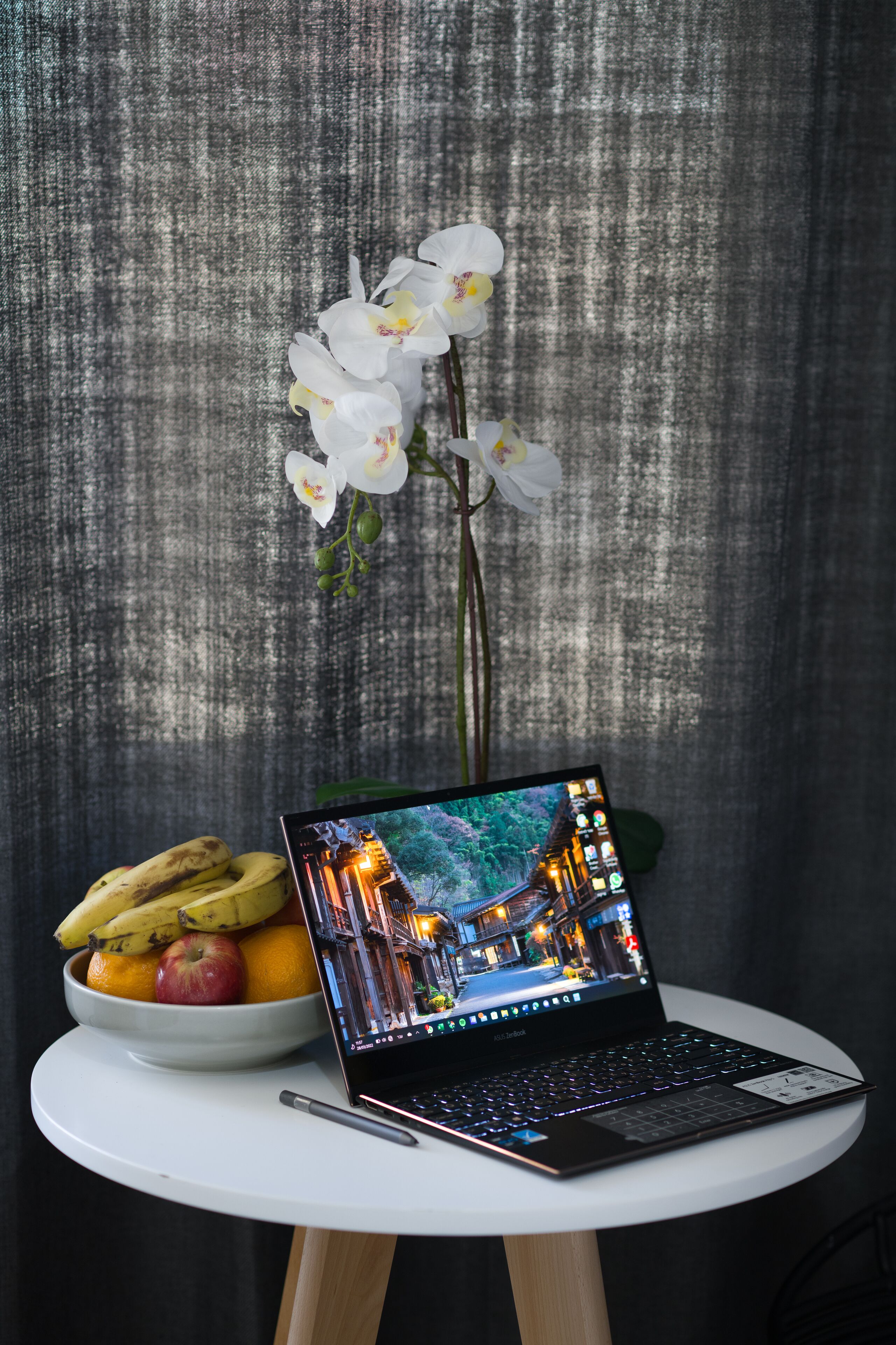 A laptop on a white table with a bowl of fruit and an orchid.