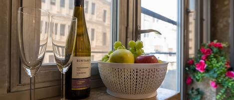 A bottle of wine, two glasses, and a bowl of fruit on a window sill.