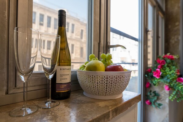 A bottle of wine, two glasses, and a bowl of fruit on a window sill.