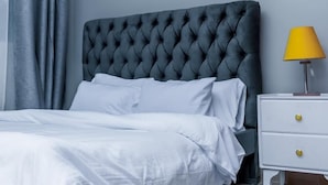 Un dormitorio con cabecera capitonada, ropa de cama blanca, una mesita de noche con cajones, una lámpara y una cortina gris.