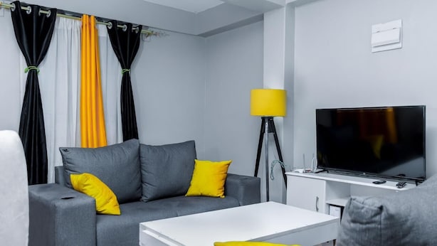 Una sala de estar moderna con un sofá gris, cojines amarillos, una mesa de centro blanca, un televisor de pantalla plana y una lámpara de pie.