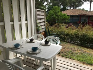 A white outdoor table with chairs, a wooden deck, and a garden with bushes and a building in the background.
